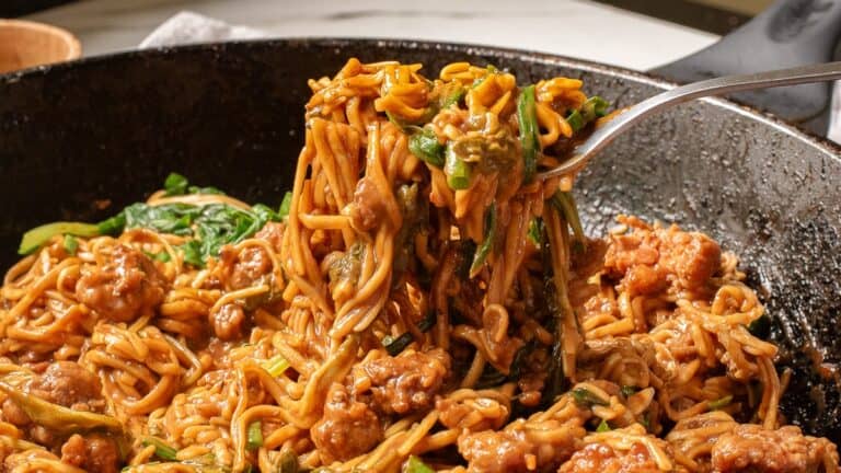 A fork lifts a serving of saucy noodles mixed with vegetables and chunks of meat from a black pan.