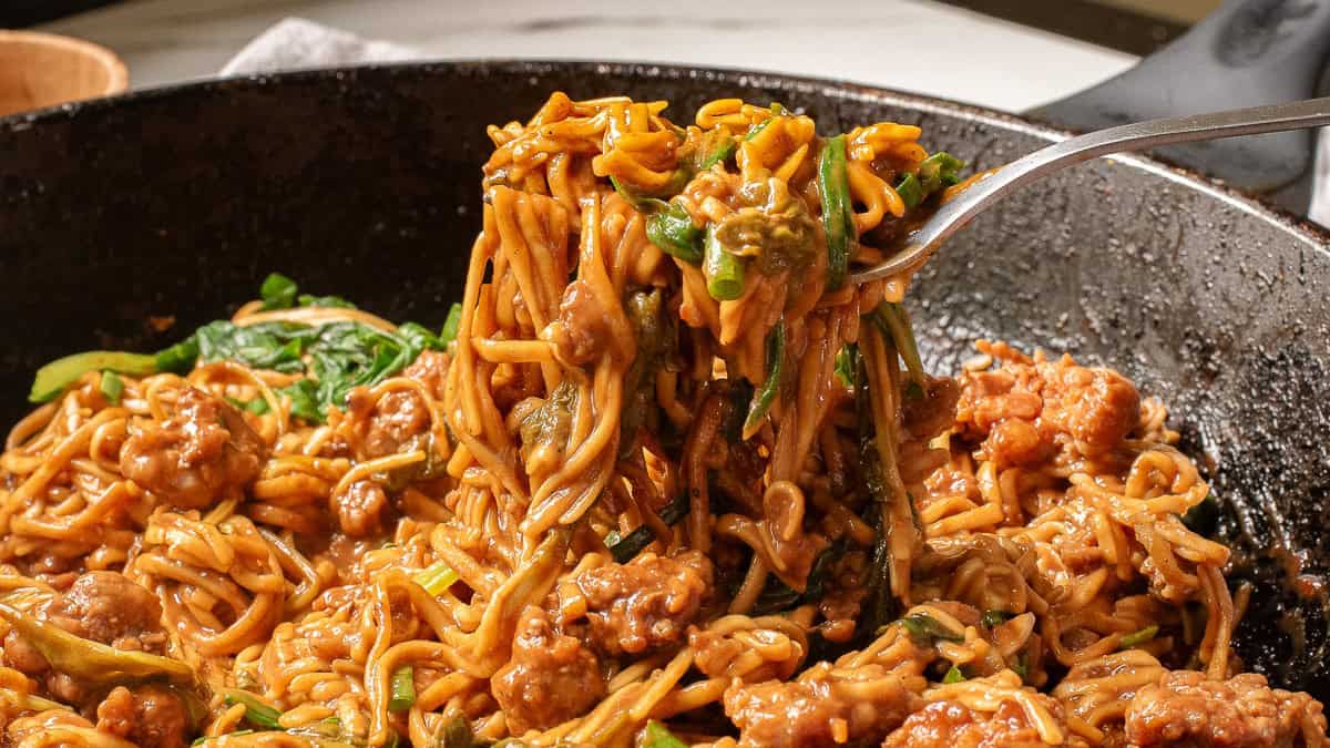 A fork lifts a tantalizing forkful of noodles mixed with sausage and vegetables from a black skillet. The dish appears to be creamy and richly seasoned, reminiscent of dan dan noodles, with visible bits of meat and greens intertwined with the noodles.