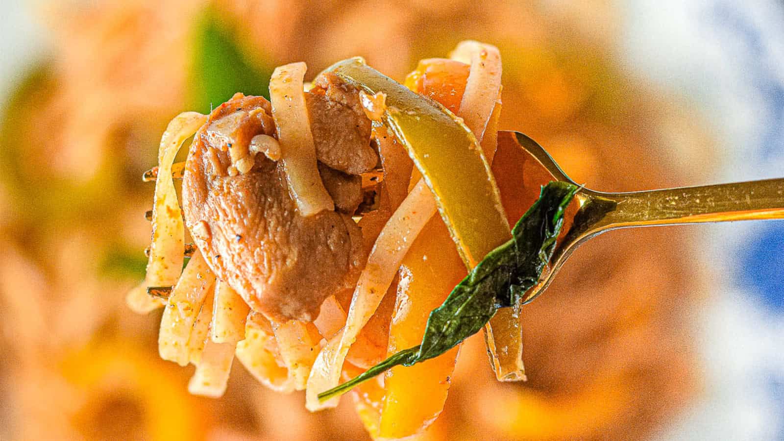 A close-up of a fork holding a portion of pasta with pieces of meat, bell pepper slices, and a basil leaf. The background is blurred, focusing on the vibrant colors and textures of the food.