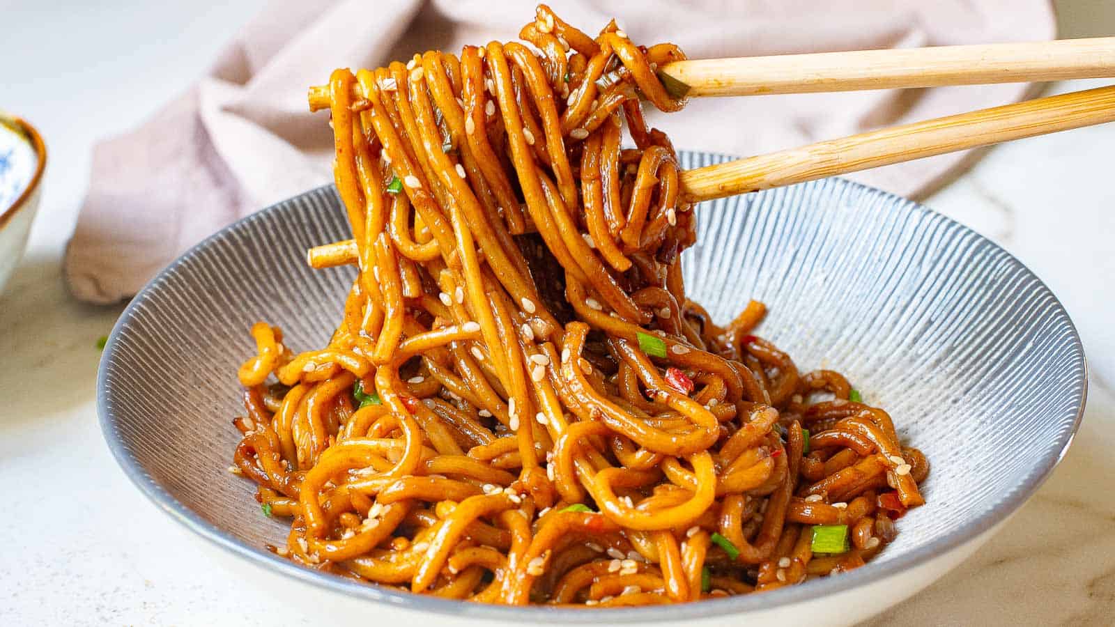 A close-up of a hand using chopsticks to lift cooked noodles from a black pan. The noodles are mixed with vegetables and sauce. A small dish is visible in the background.