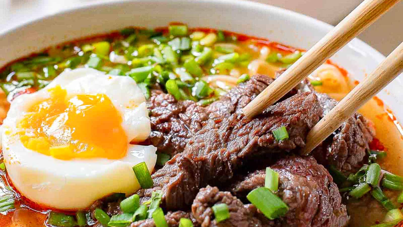 Close-up of a bowl of ramen with rich broth, topped with slices of tender beef, a perfectly poached egg with a runny yolk, and garnished with chopped green onions. Chopsticks are holding up a piece of beef.