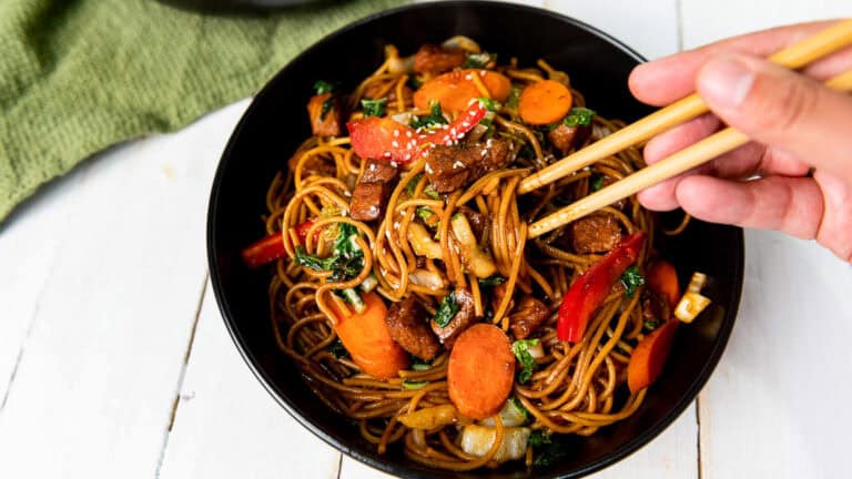 A hand using chopsticks to hold a noodle dish with vegetables and meat in a black bowl on a white wooden surface.