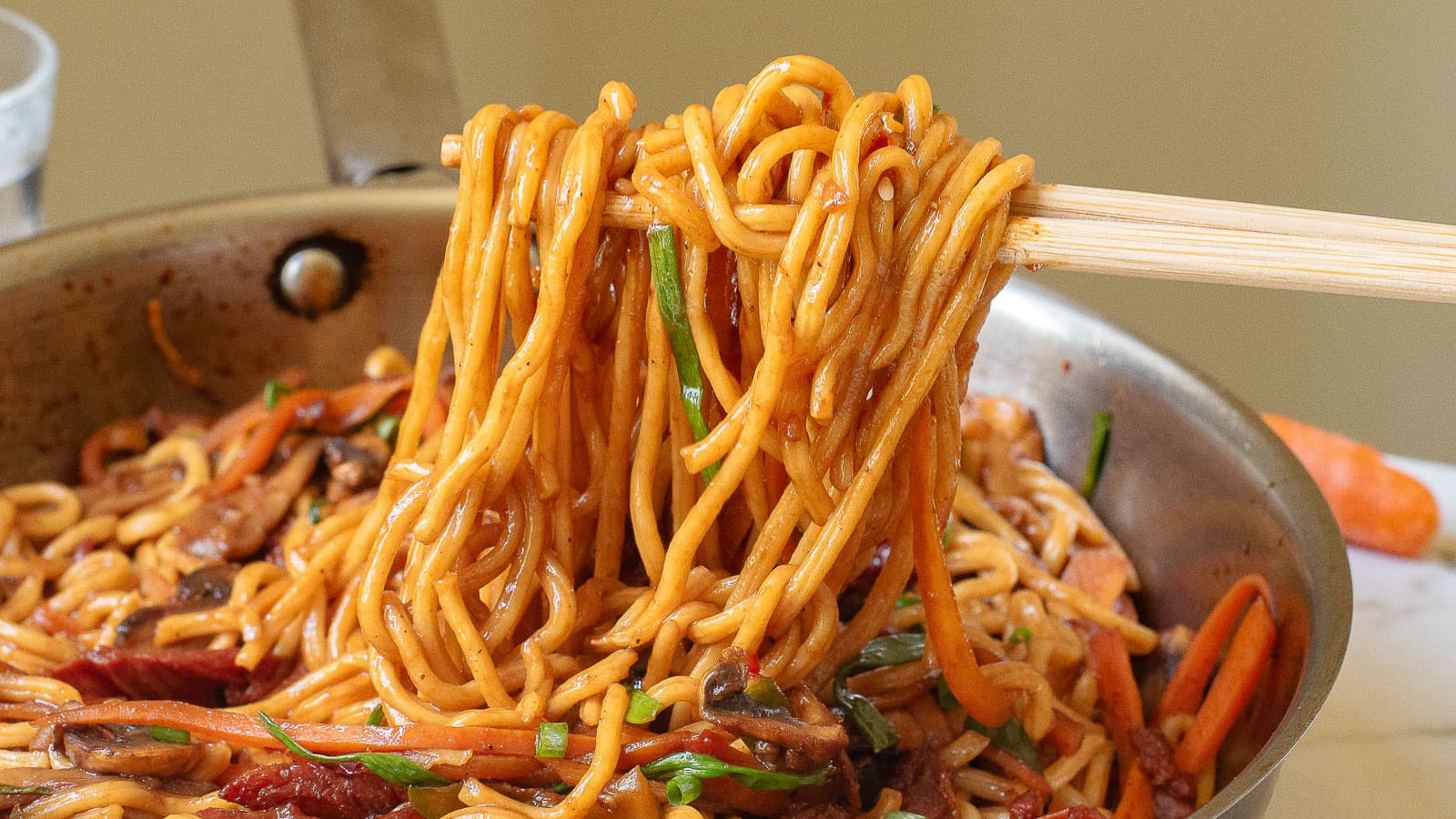 A bowl of saucy noodles is being lifted with chopsticks over a marble countertop. A carrot is partially visible in the background.