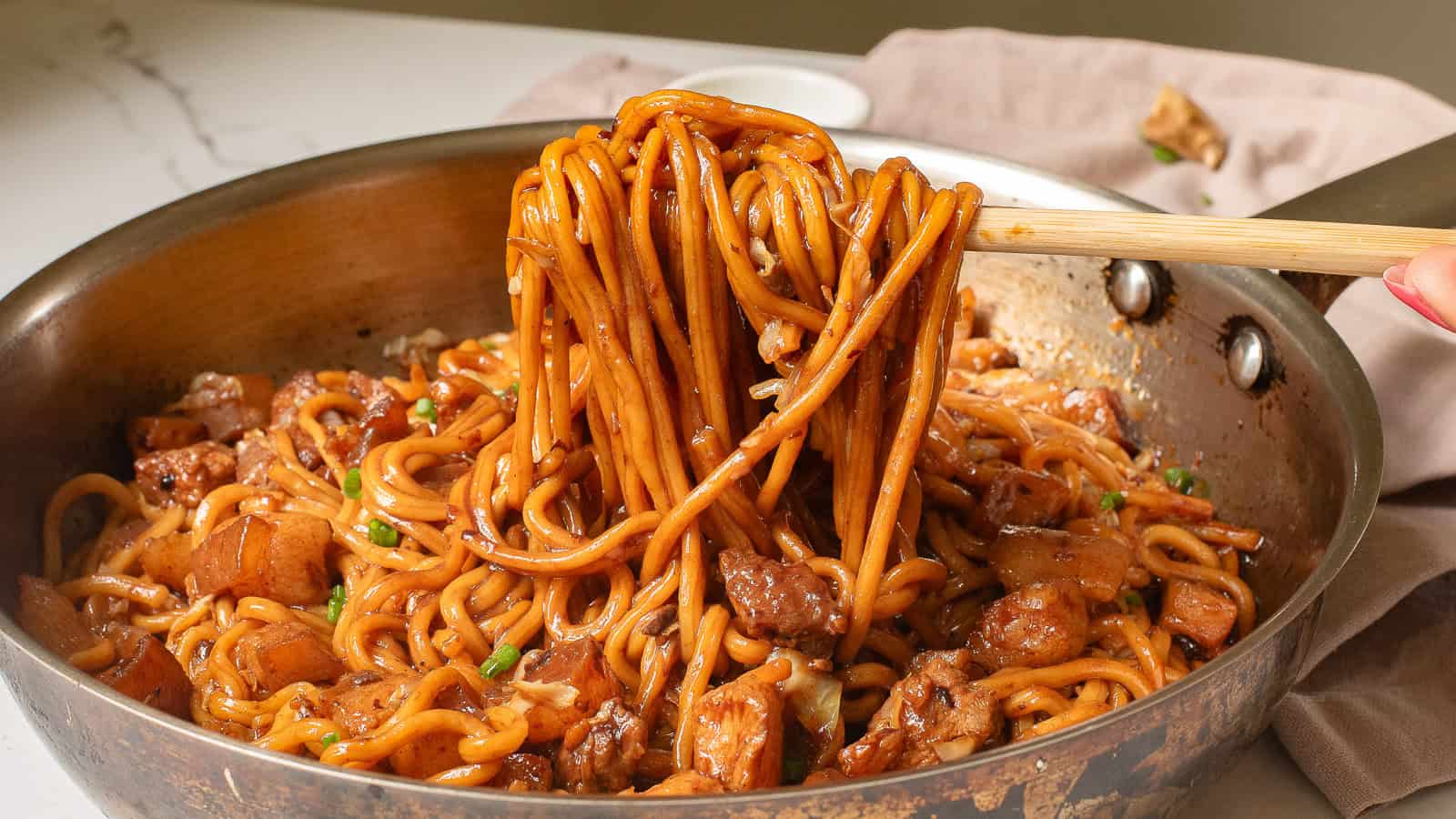 A close-up of a pan filled with savory noodles and chunks of meat being lifted by chopsticks. The dish appears well-seasoned with sauce, and green garnishes are sprinkled on top. A beige napkin and a piece of dumpling are in the background.