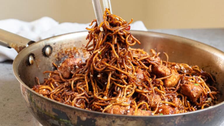 Stir-fried noodles with chunks of meat are being mixed in a pan using chopsticks.