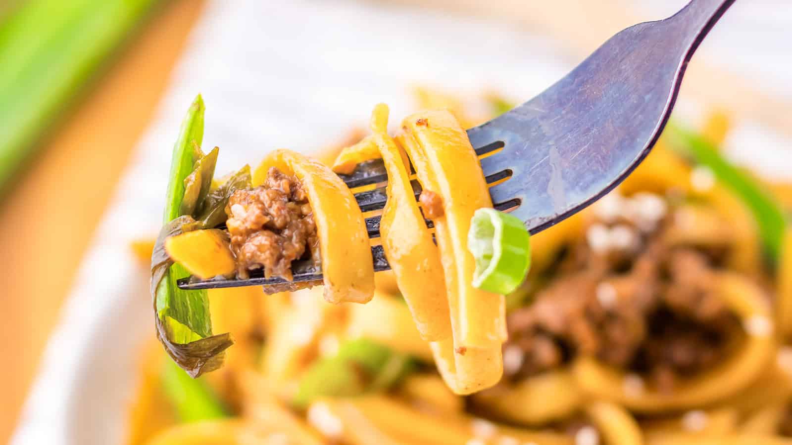 A close-up of a fork holding a bite of pasta with ground meat, green onions, and sauce. The pasta is wrapped around the fork tines, displaying the dish's ingredients and textures in detail, with a blurred background featuring more of the meal.