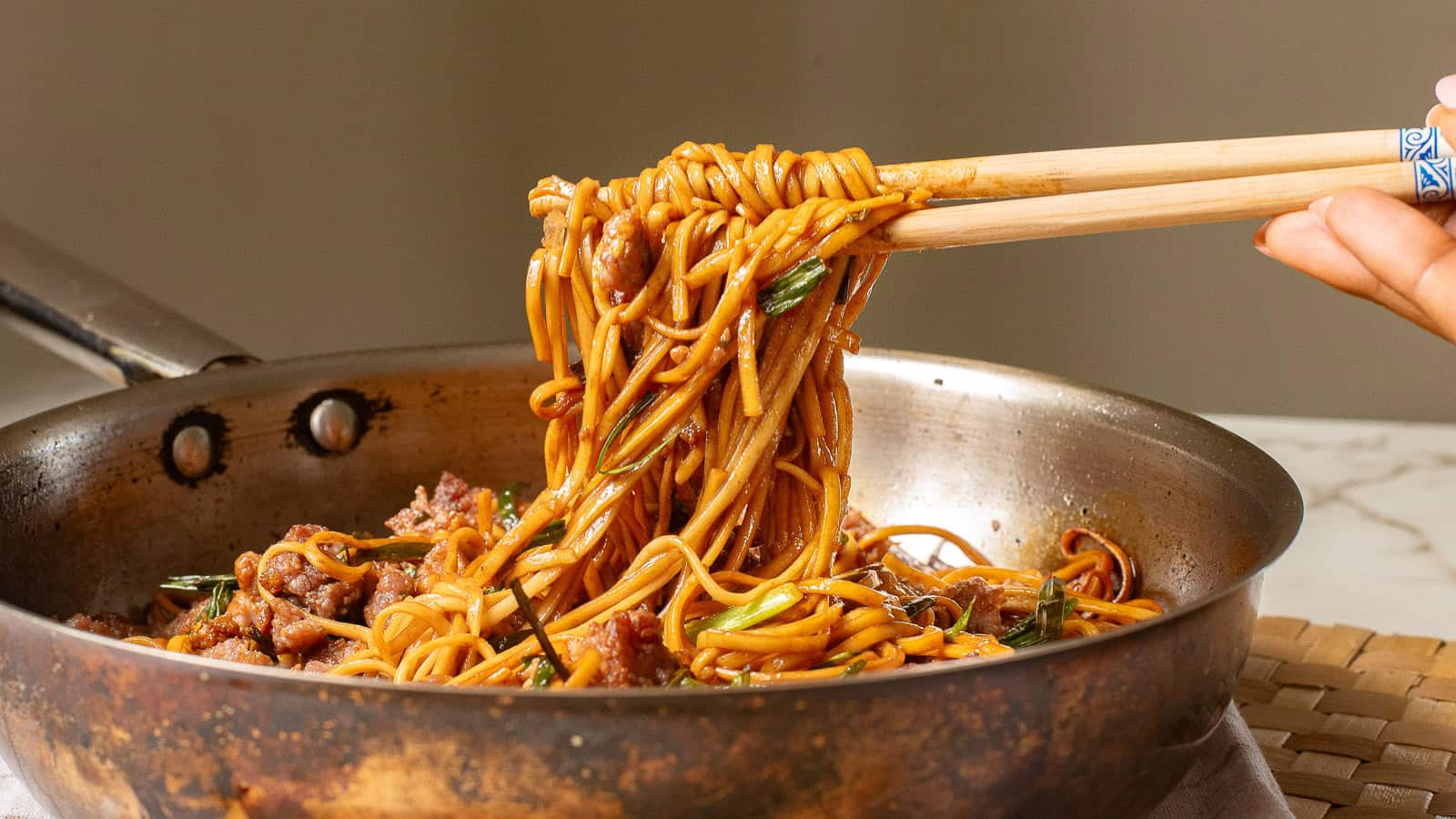 A close-up of a pan filled with stir-fried noodles. A pair of chopsticks is lifting a portion of the noodles, which are mixed with small pieces of meat and green vegetables. The dish appears appetizing and is set on a woven mat.