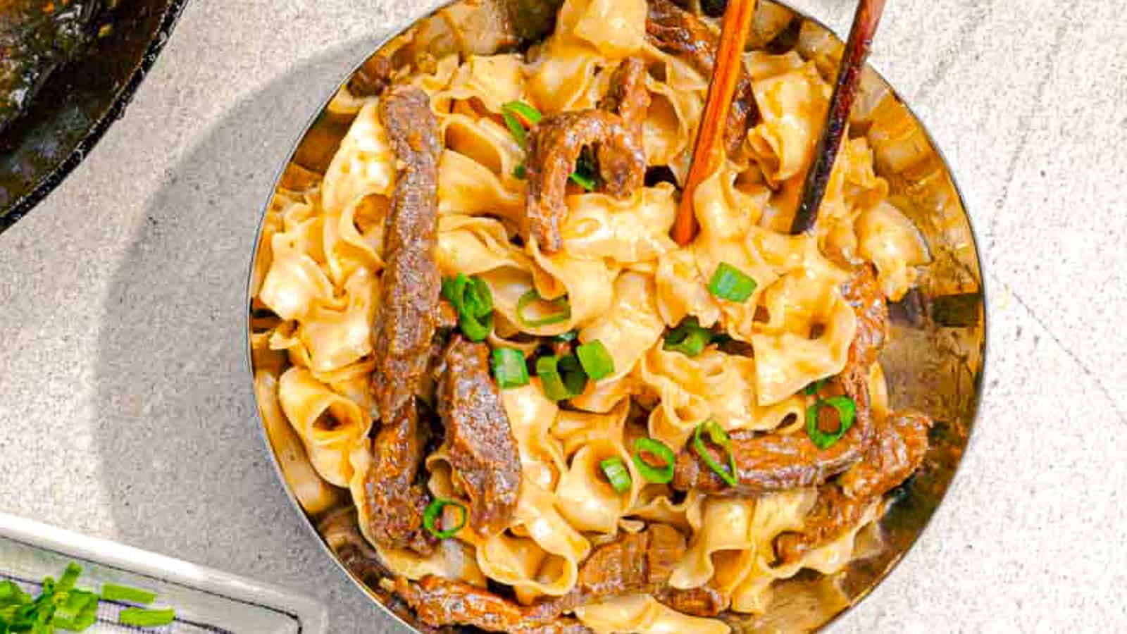 A bowl of ribbon noodles with sliced beef, topped with chopped green onions, and a pair of chopsticks.