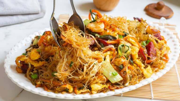 A plate of stir-fried glass noodles with vegetables, shrimp, and eggs, on a bamboo mat.