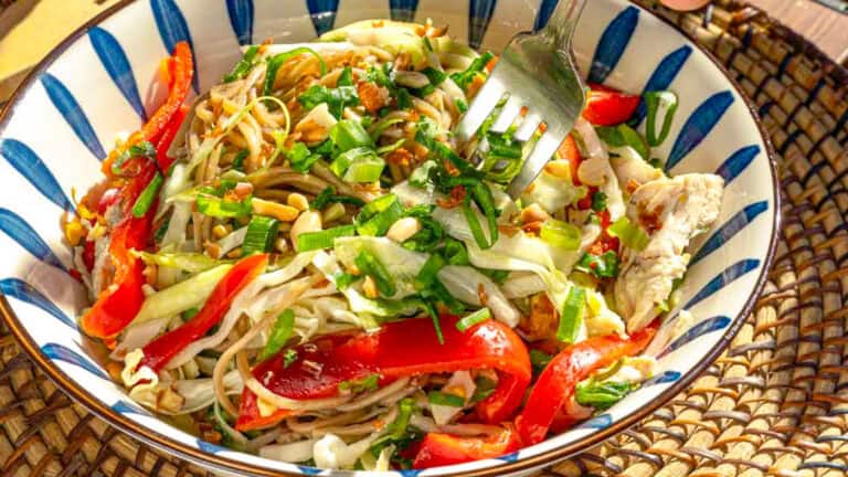 A bowl of noodle salad with red peppers, cabbage, chopped greens, and peanuts, with a fork poised over the dish.