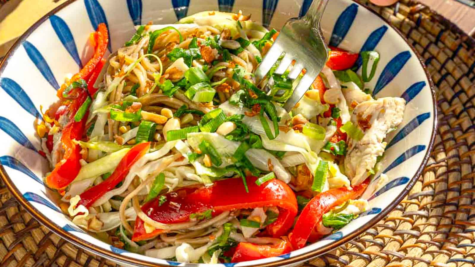 A colorful salad in a striped bowl, featuring mixed vegetables such as red bell peppers and shredded cabbage, garnished with crushed nuts and fresh herbs. A fork is seen lifting a portion, emphasizing the dish's vibrant, fresh ingredients.