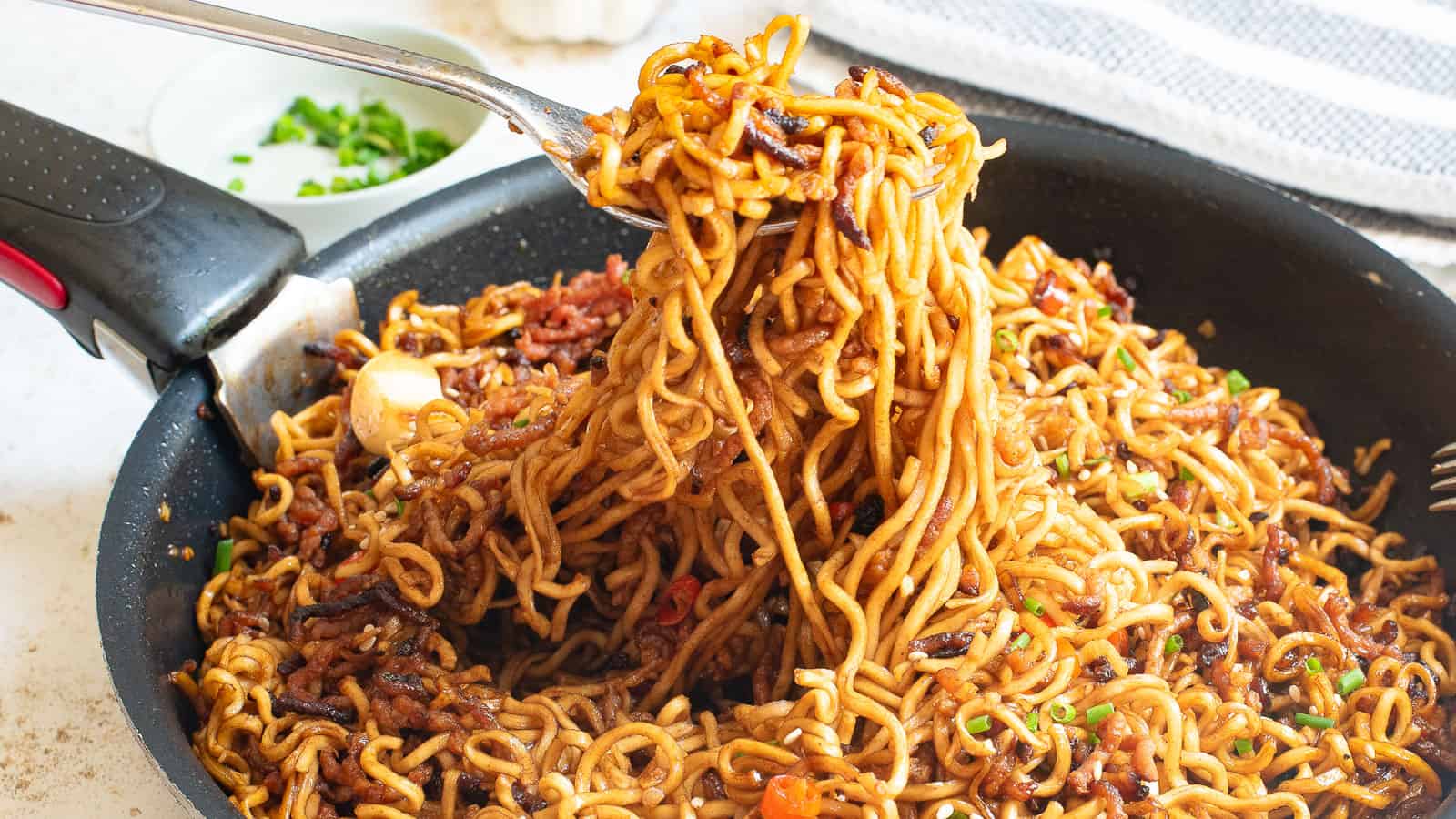 A fork lifting a portion of cooked noodles with vegetables from a pan, with a bowl of chopped green onions in the background.