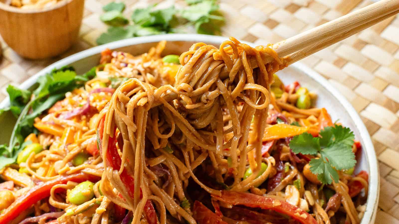 Chopsticks holding a portion of spicy peanut noodles with assorted vegetables and cilantro on a woven mat background.