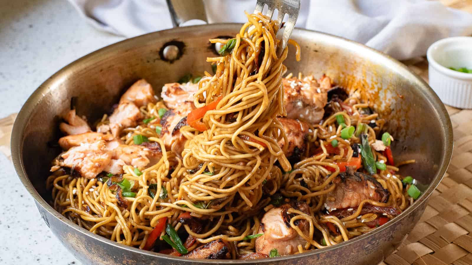 Stir-fried noodles with vegetables and chunks of salmon in a skillet, with a fork lifting a portion.