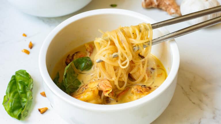 A bowl of soup with noodles, chunks of chicken, and basil leaves, with chopsticks lifting noodles. Pieces of garlic and ginger are scattered around the bowl on a marble surface.