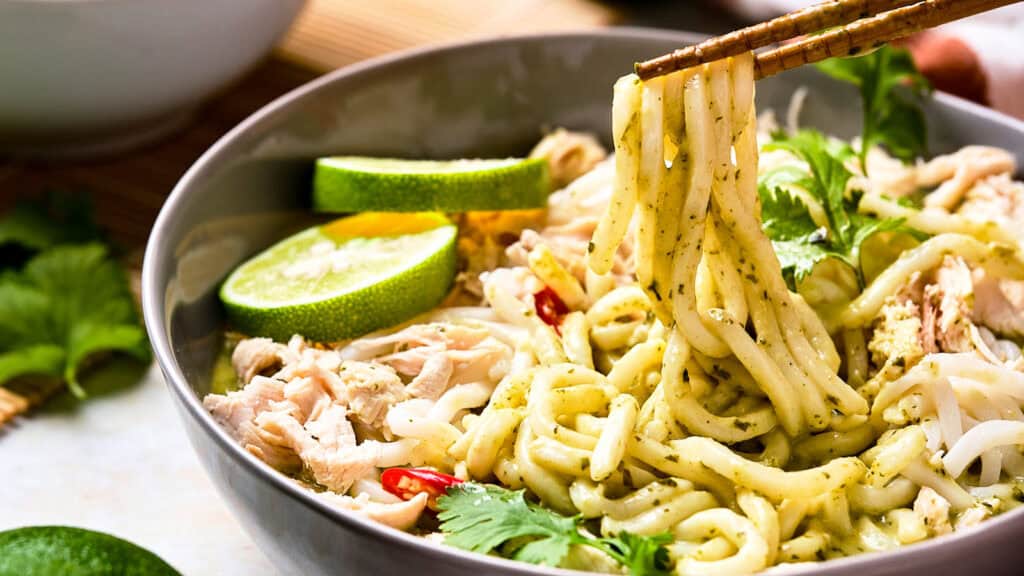 A bowl of noodles with green sauce, shredded chicken, lime slices, cilantro, and red chili, with chopsticks lifting noodles.