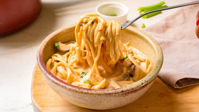 A bowl of creamy pasta with mushrooms and herbs, being twirled on a fork, set on a wooden board with a small sauce dish in the background.