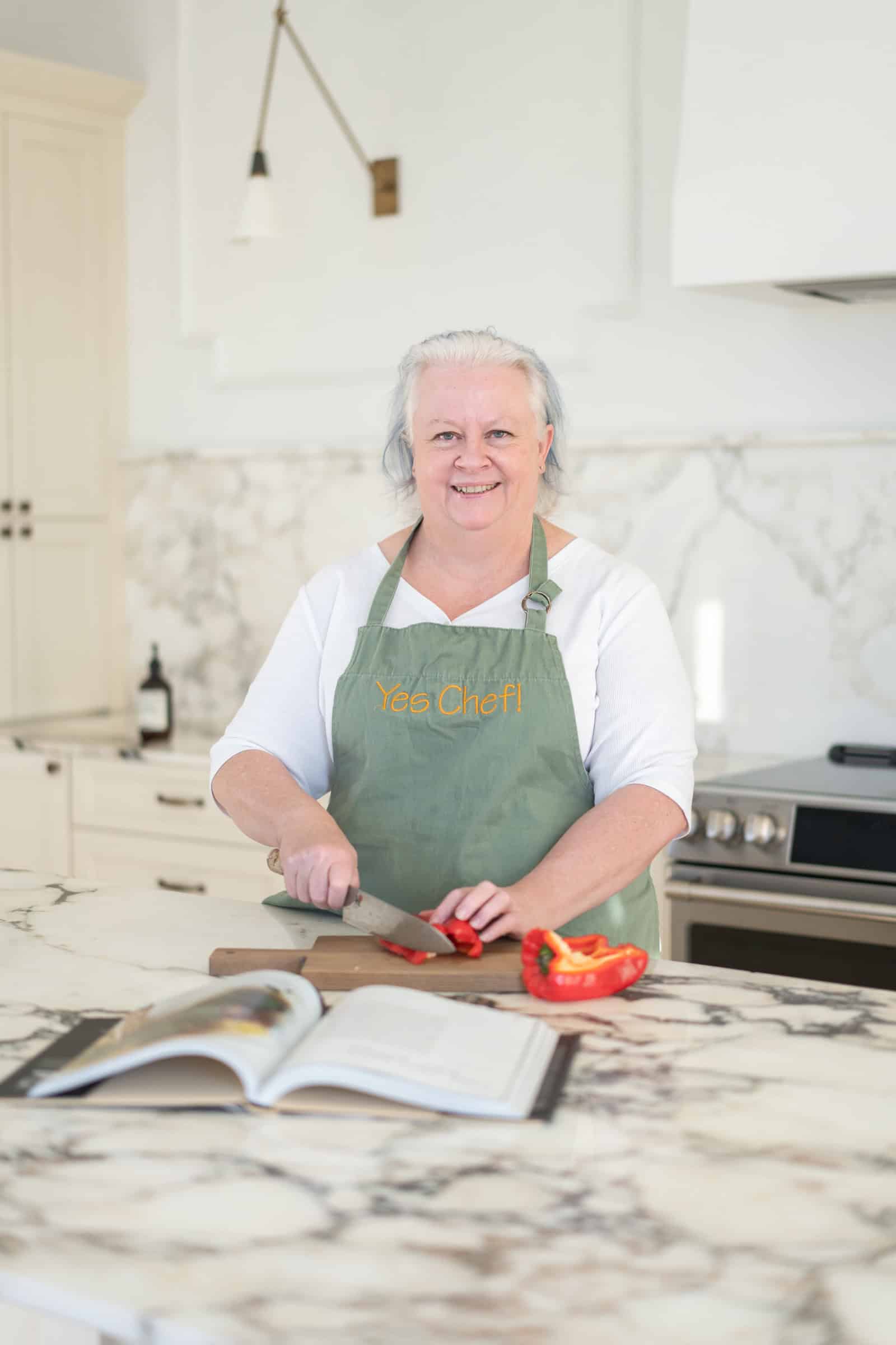 In a cozy kitchen, a person skillfully slices a red bell pepper on the chopping board, with an open cookbook in the foreground. It's moments like these that tell our own 'About Us' story through every cherished recipe page turned.