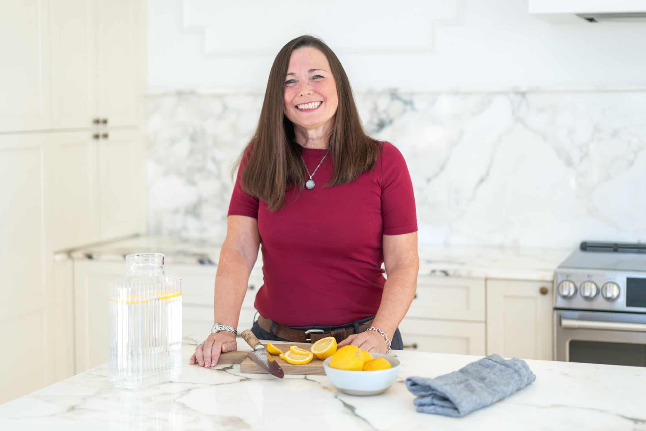 In the heart of our kitchen, a woman in a red shirt smiles warmly. On the marble counter, a cutting board holds sliced oranges beside a knife and bowl of fruit. Nearby, a towel and water pitcher await use. This scene captures the essence of our passion for fresh ingredients at About Us.