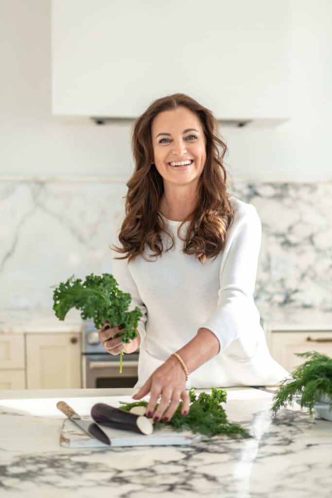 In a bright, white kitchen, the woman smiles warmly as she chops fresh vegetables on a marble countertop, embodying the spirit of our About Us story.